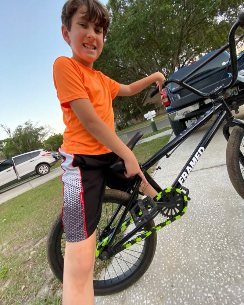 Child in orange shirt and black shorts standing next to a bicycle on a street and showing off his bright green and black Insane Chains Caps