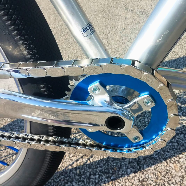 Close-up of a bicycle chainring with blue anodized finish and chrome insane chains snapped onto the chain, on a gravel surface